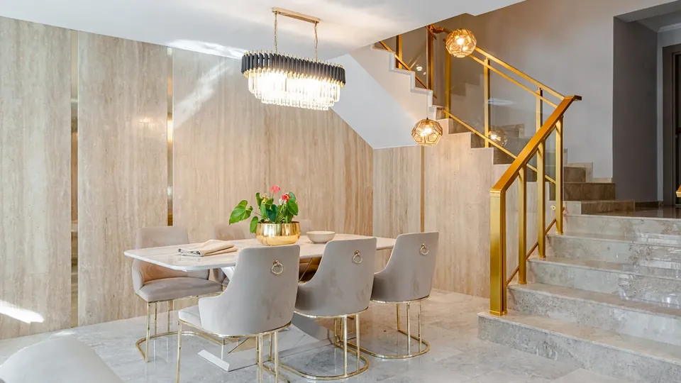 Dining room with a warm travertine-look panel wall with champagne inlays, a marble dining table and a gold staircase railing